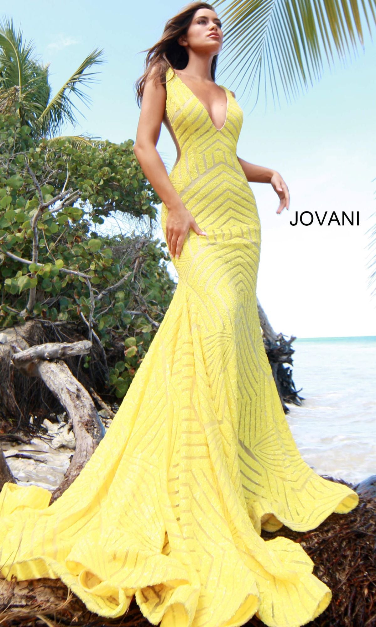 A woman stands on a beach in a sleeveless, floor-length yellow Jovani Plus-Size Mermaid Prom Dress with sequins and geometric patterns, with the ocean, greenery, and palm trees in the background.